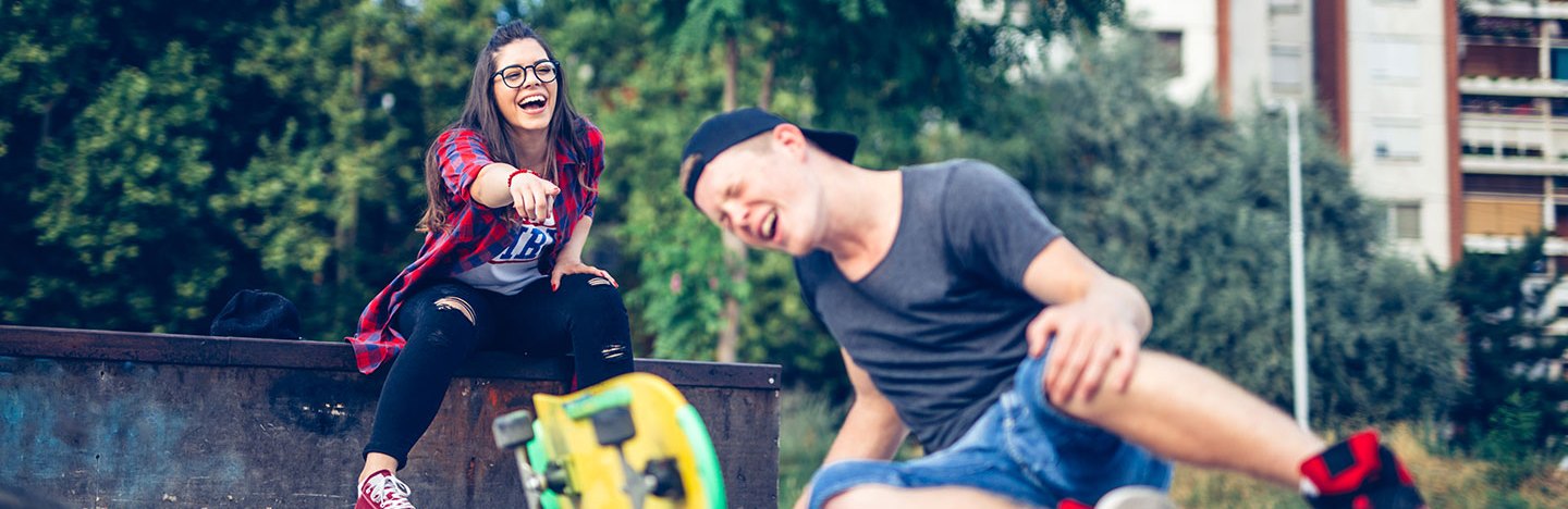 a pale teen boy has fallen off his skateboard and seems injured, behind him a teen girl points and laughs at him