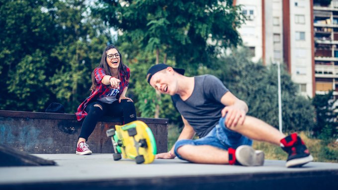 a pale teen boy has fallen off his skateboard and seems injured, behind him a teen girl points and laughs at him