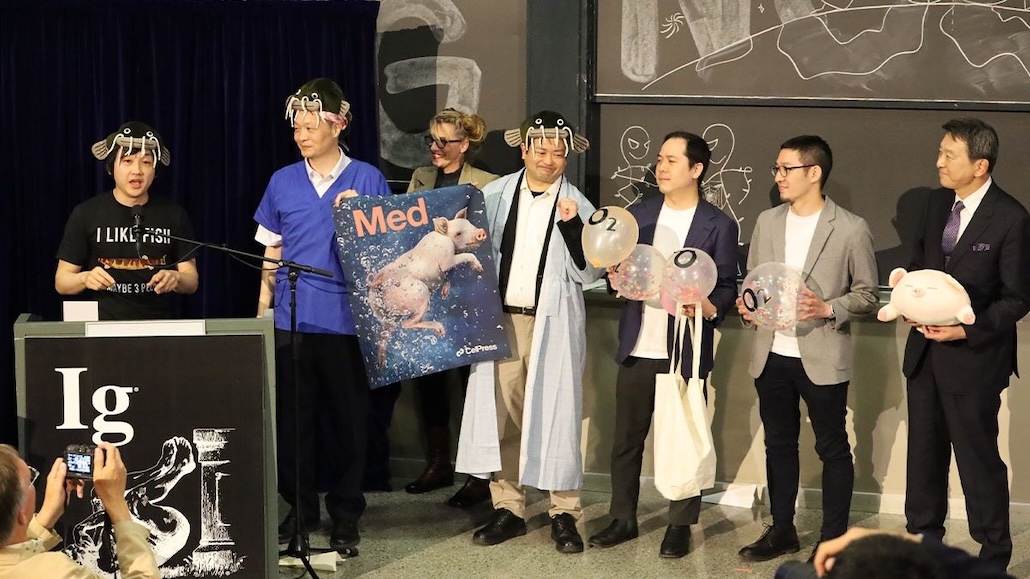 a group of men stand on stage, some holding balloons labeled "O2" for oxygen, some holding stuffed pigs, and the one at the podium wearing a fish-shaped hat