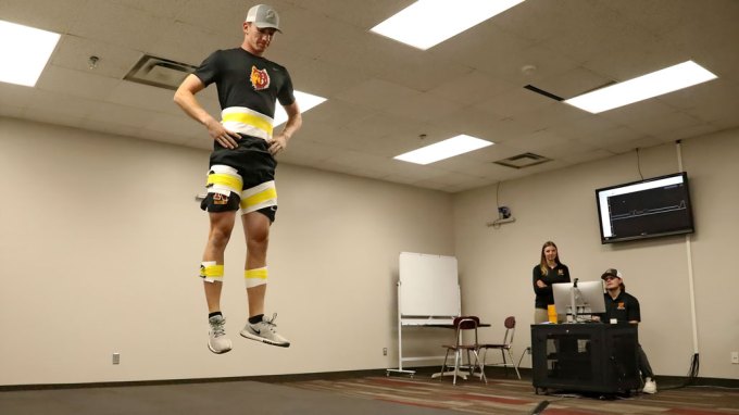 A man in sports attire jumps in a lap. A whiteboard sits in the corner. A sits at a desk with a computer next to the wall. Jessica Talmage stands next to him. She is a white woman with long blonde hair.