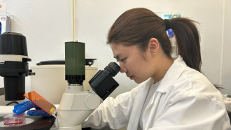 A teen in a lab coat looks into a microscope. A lab flask containing red liquid and cells to model a form of arthritis sits on the microscope stage.