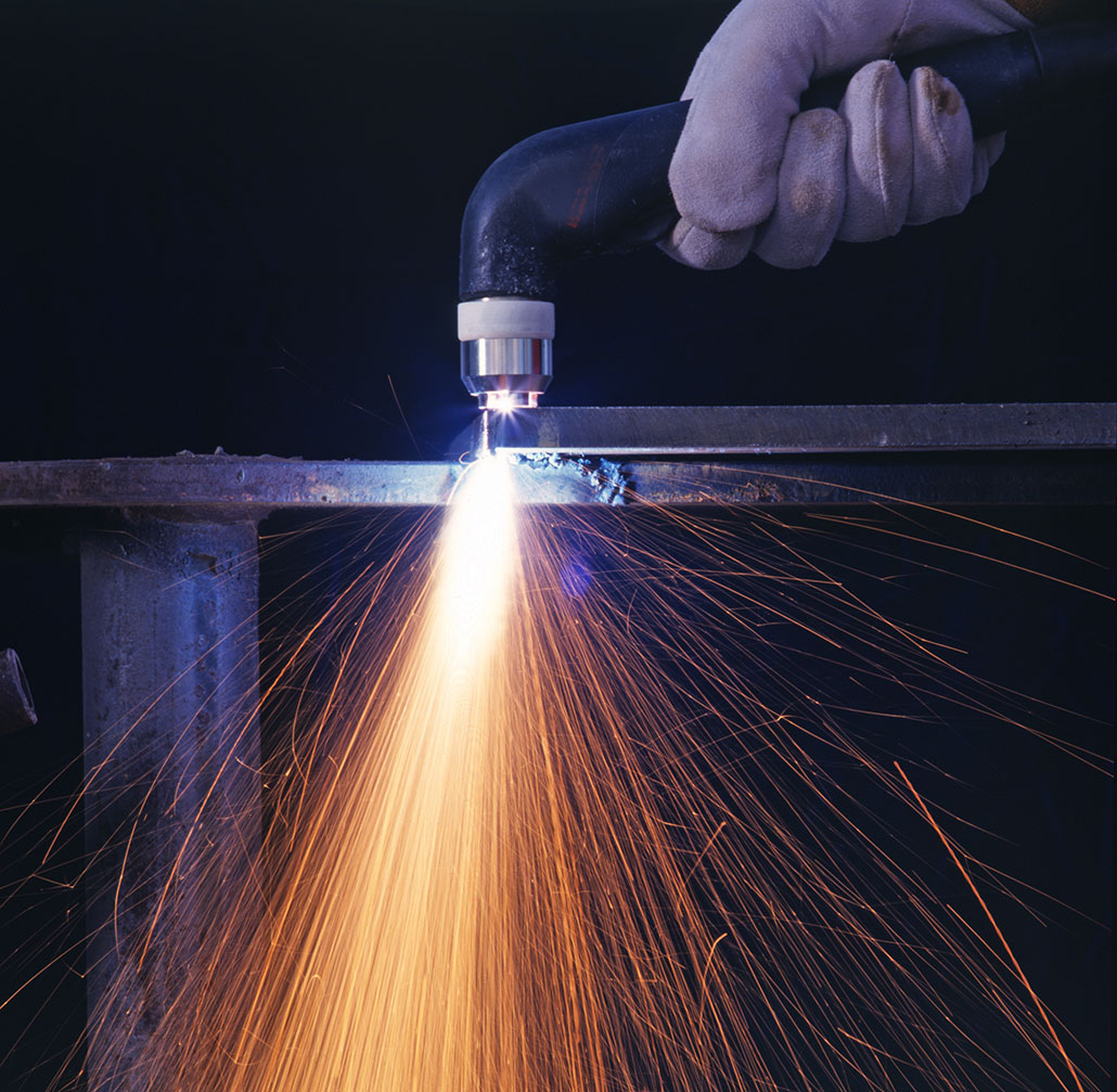 A gloved hand can be seen holding a plasma torch to a sheet of metal. Sparks fly as the torch cuts.