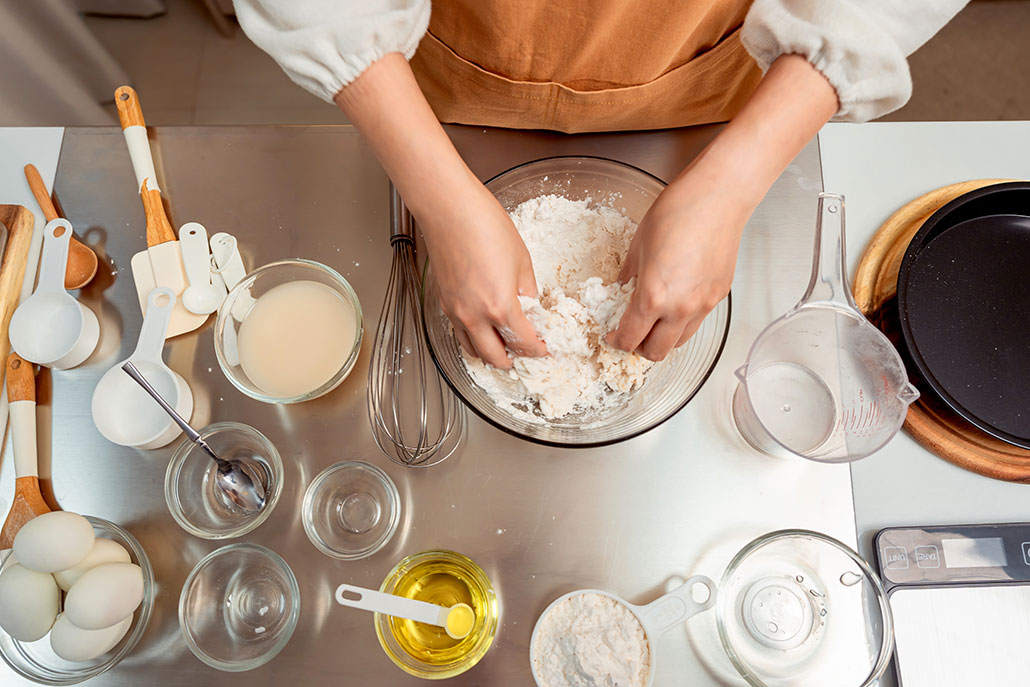 a top down picture of a countertop where someone is making dough, bowls of ingredients and utensils are scattered around the mixing bowl (which is full of dough being mixed)