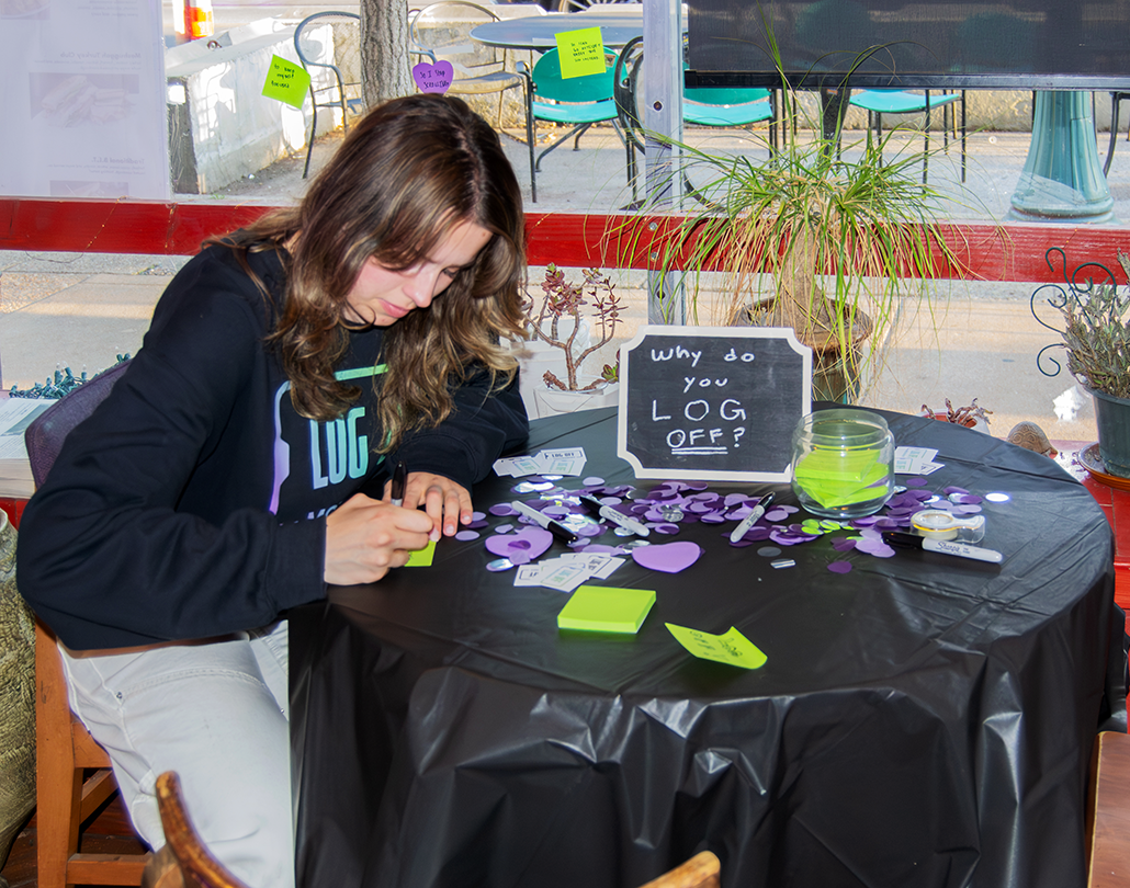 Emma Lembke, a young white woman with long dark brown hair, sitas at a round table and writes down reasons why she logs off. A sign on the table says 'Why do you LOG OFF?' There are sticky notes and markers arranged around the table.