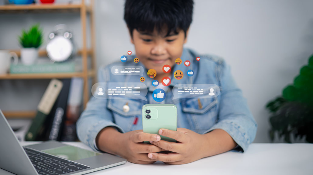 a young Asian kid on a phone, social media icons float around to illustrate his participationon on social media