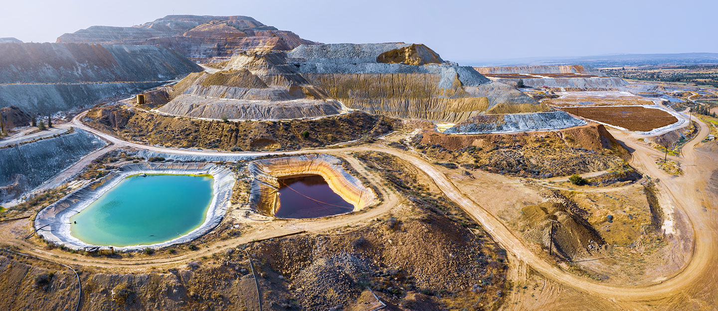 an aerial view of a mining operation, in the middle is a hill that has been mined, in the bottom of the image are large water ponds of different colors. One is bright blue-green and the other is ringed in orange with dark red tinted water.