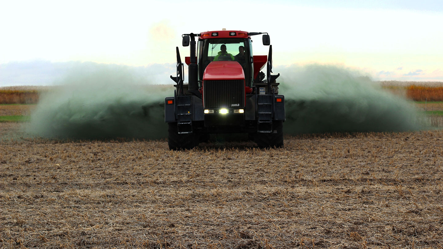 a red tractor drives toward the viewer over an empty field. It is spraying basalt on the field, behind and to the sides of the tractor.