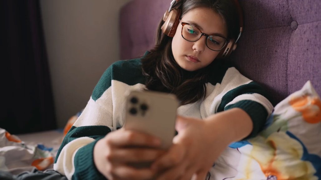 a still frame of a girl wearing headphones on her bed scrolling through a smart phone