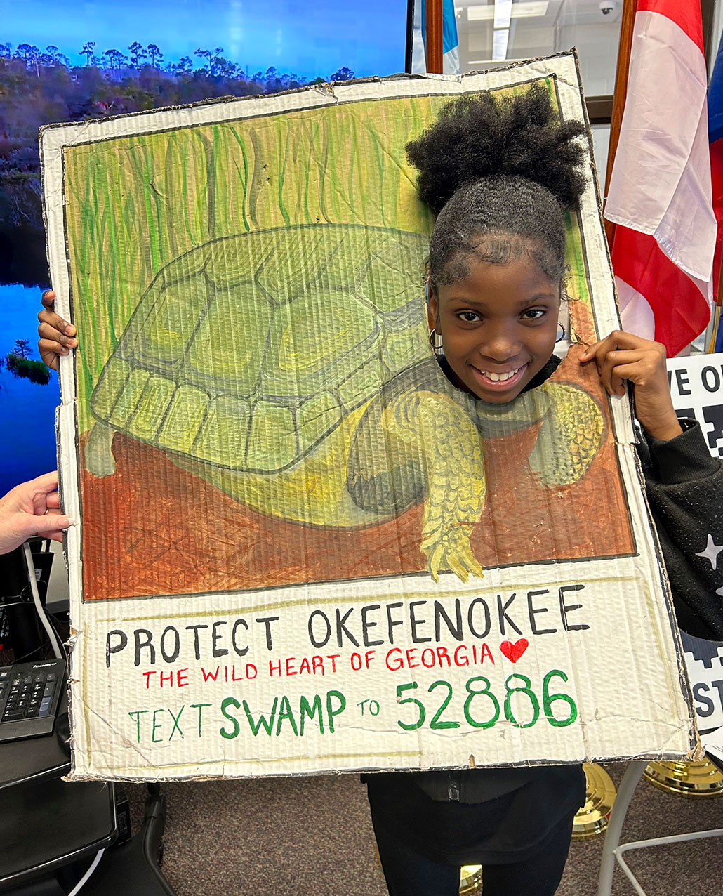 a smiling young Black girl puts her head through a sign where a tortoise's head would be. The bottom of the sign reads 'Protect Okefenokee, The Wild Heart of Georgia' Text SWAMP to 52886