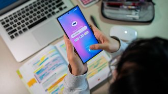 a top down view of a girl with dark hair sitting at a desk studying, she has a notebook full of highlighted notes open in front of her laptop, a pencil case is in the top right corner, and she is using her phone to access a chatbot