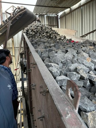 a chute runs diagonally from the bottom right to the top middle of the image. It is full of large chunks of grey rock. A worker stands to the side supervising.