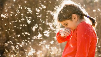 A young girl standing outside is surrounded by floating plant fluff and pollen. She's sneezing.