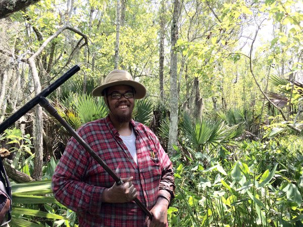 Elliott White Jr. stands in front of thick shrubbery. He's a Black man with a black beard and brown eyes. He's wearing a wide-brimmed hat, eyeglasses and a red flannel shirt
