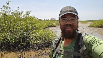 Elliot White stands in front of a wetlands. He's a Black man wearing a cap, eyeglasses and green tee-shirt. He has a black beard and brown eyes.