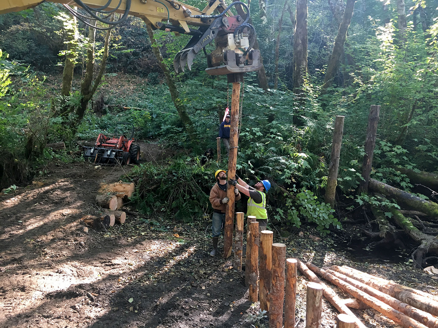Two workers are pounding a line of large logs into a creek with the help of heavy construction equipment