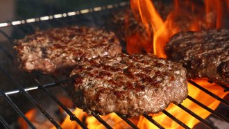 several beef hamburger patties cooking on a grill, with flames shoot up in the background