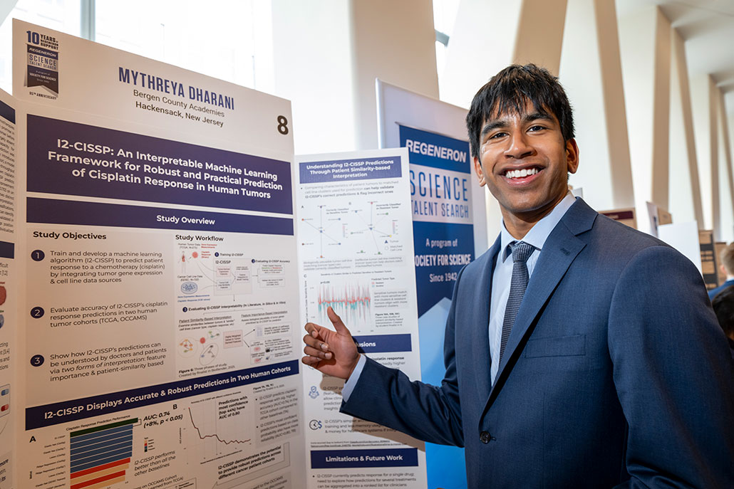 a photo of a young man with brown skin and dark hair beaming as he stands in front of his research project