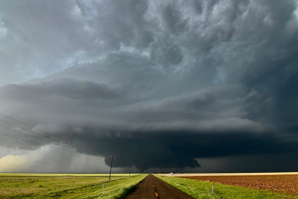 a photo of a dark sky and flat landscape. A straight road in the middle of the phot leads to a rotating stomr and tornado touching down on the horizon