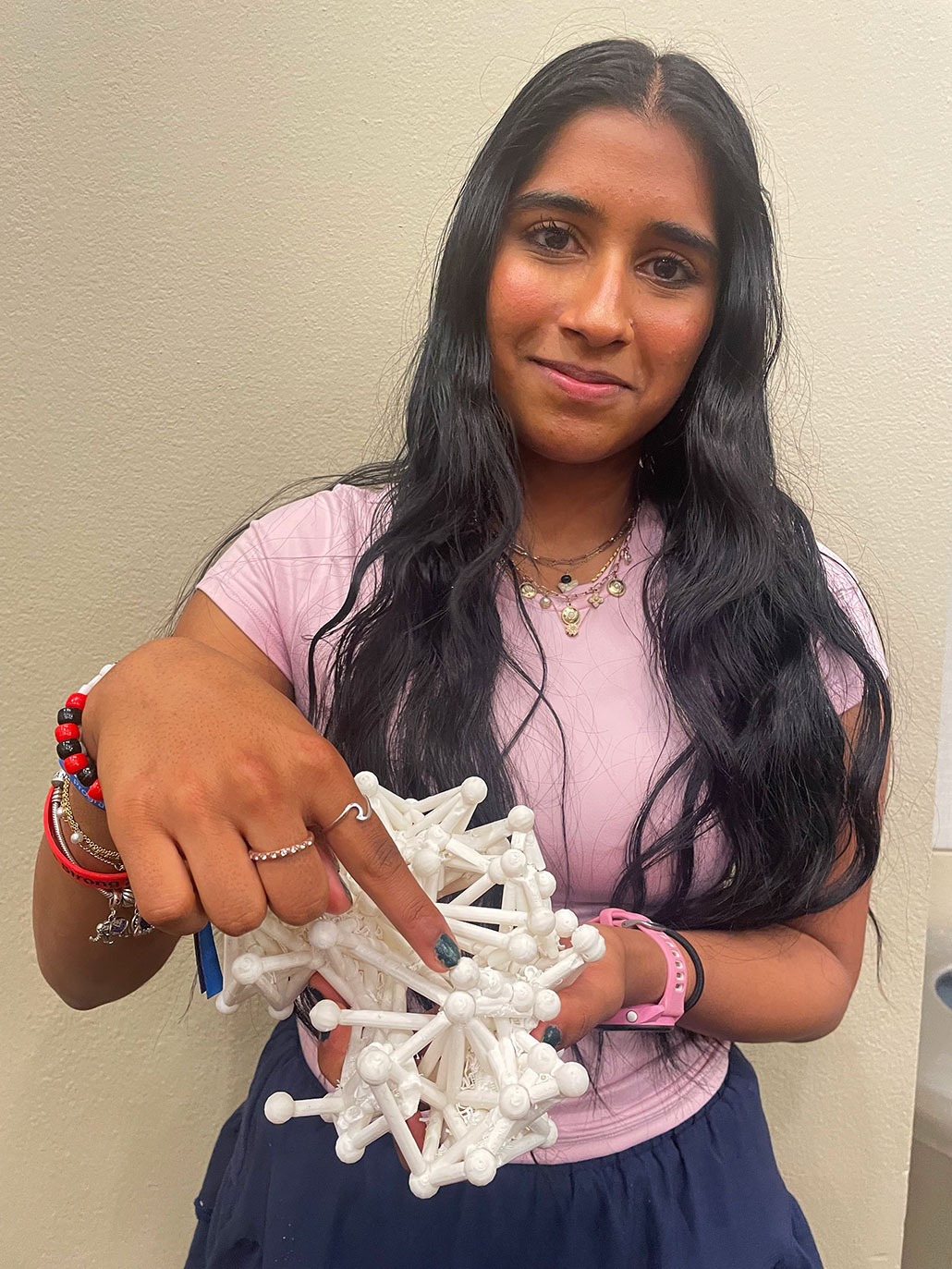 Sowmya Sankaran, a young woman in a pink top, holds a white 3D-printed geometric molecular structure toward the camera.