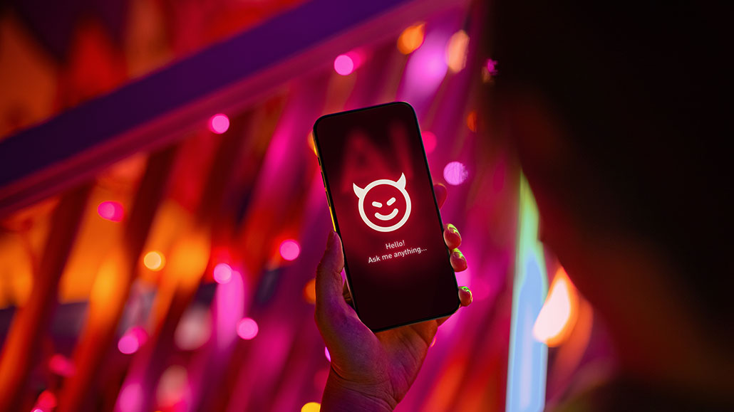 A hand holds a smartphone displaying an AI chatbot interface with a mischievous devil emoji and the prompt 'Hello! Ask me anything…' against a backdrop of vivid pink and red neon lights.