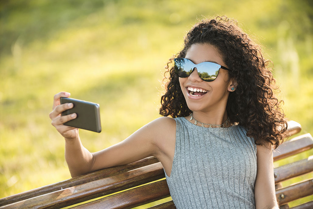 a young woman with curly brown hair and wearing sunglasses smiles for her phone while sitting on a bench