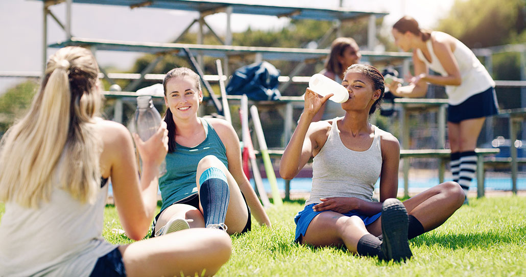 A group of female athletes in sportswear sit on grass in the sun, drinking water and sports drinks during a break from outdoor practice.