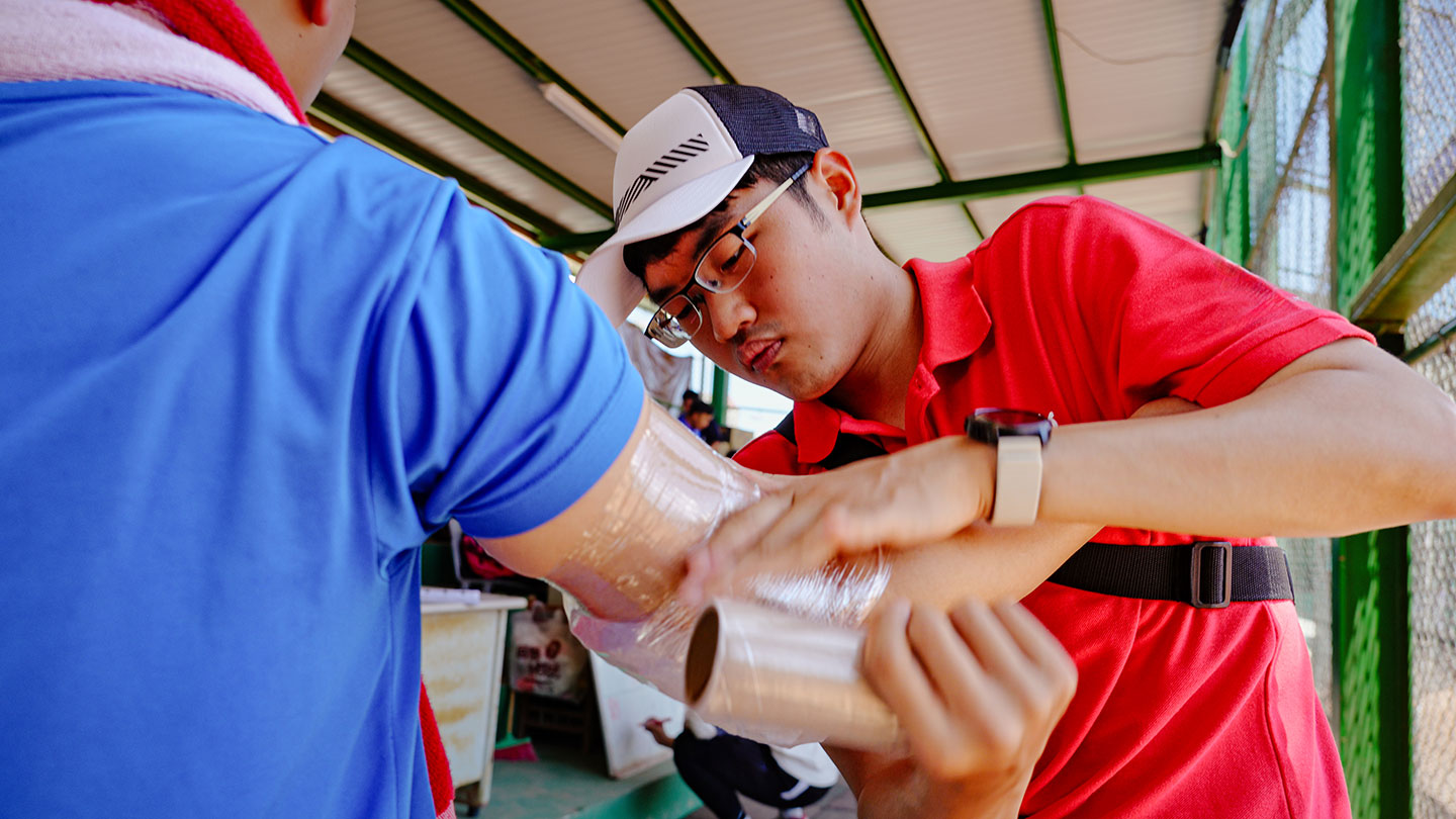 A young man in a red polo shirt and cap applies a clear plastic wrap over an ice pack to another person's forearm and elbow area at what appears to be a baseball facility, concentrating closely on the task.