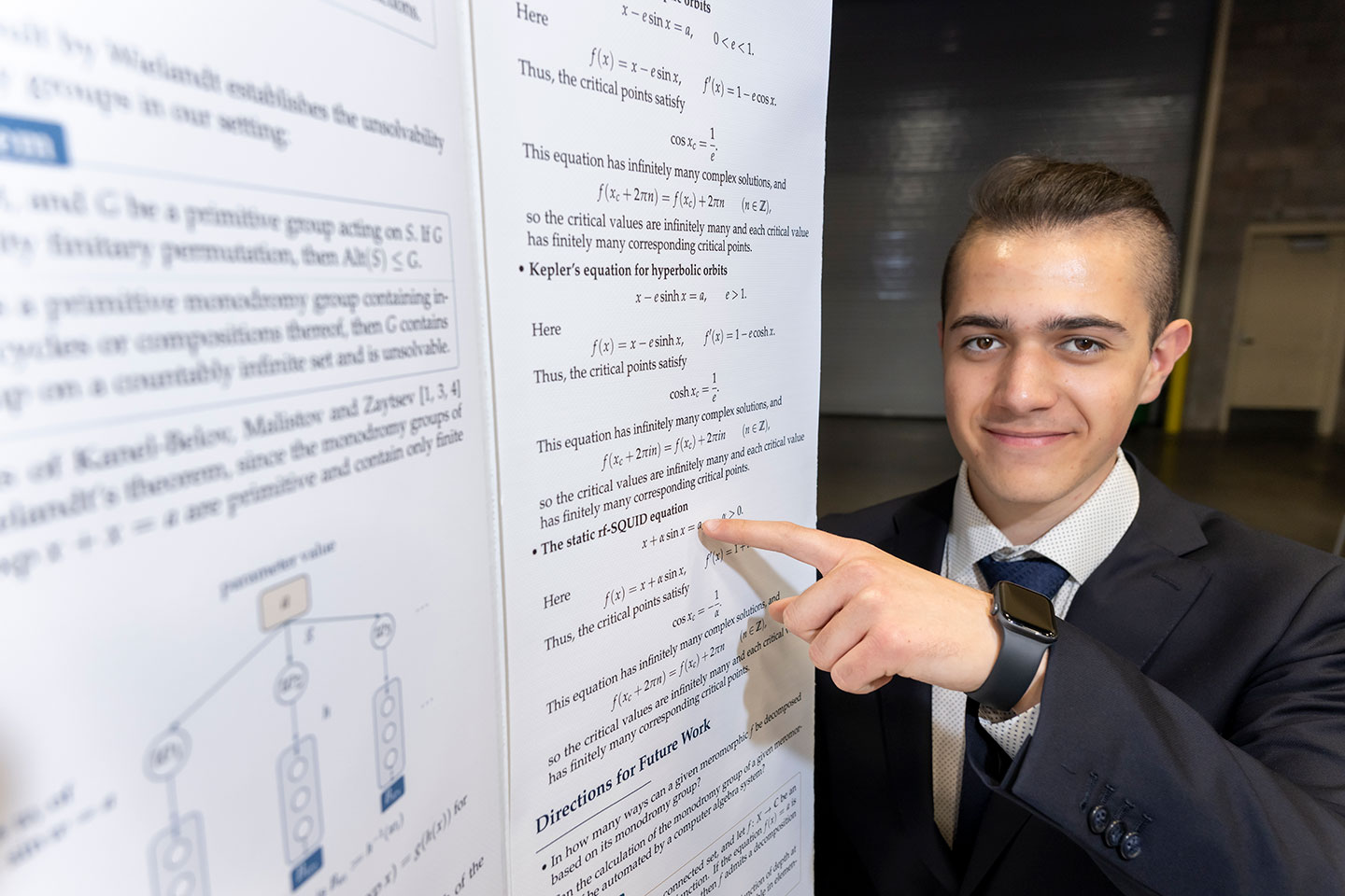 Nikola Veselinov, a teenage boy in a dark suit and tie, points to a mathematical equation on his research poster, which covers topics including Kepler's equation for hyperbolic orbits and the static rf-SQUID equation.