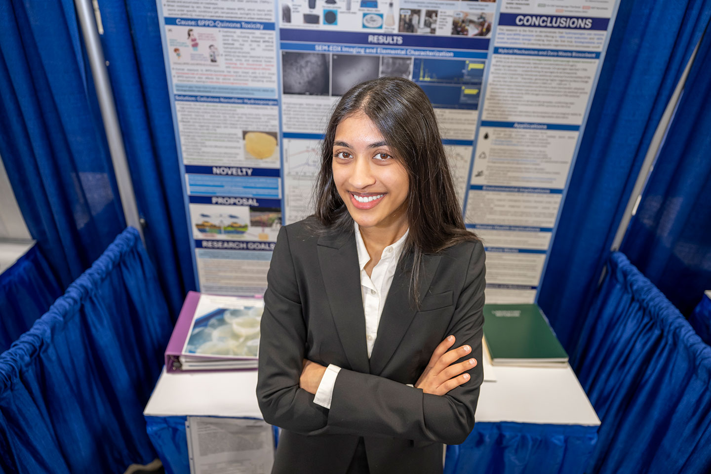 Lakshmi Agrawal, a teenage girl in a black blazer, stands with arms crossed and smiles in front of her science fair research poster on cellulose nanofiber hydrogels, with sections visible including Results, Novelty, Proposal, and Research Goals.