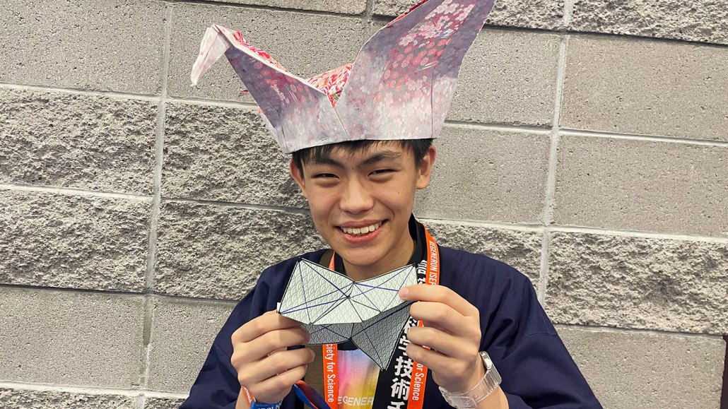 A smiling teenage boy wearing a floral origami crane as a hat holds a geometric folded paper structure, with a Regeneron ISEF lanyard around his neck, posing against a concrete block wall.
