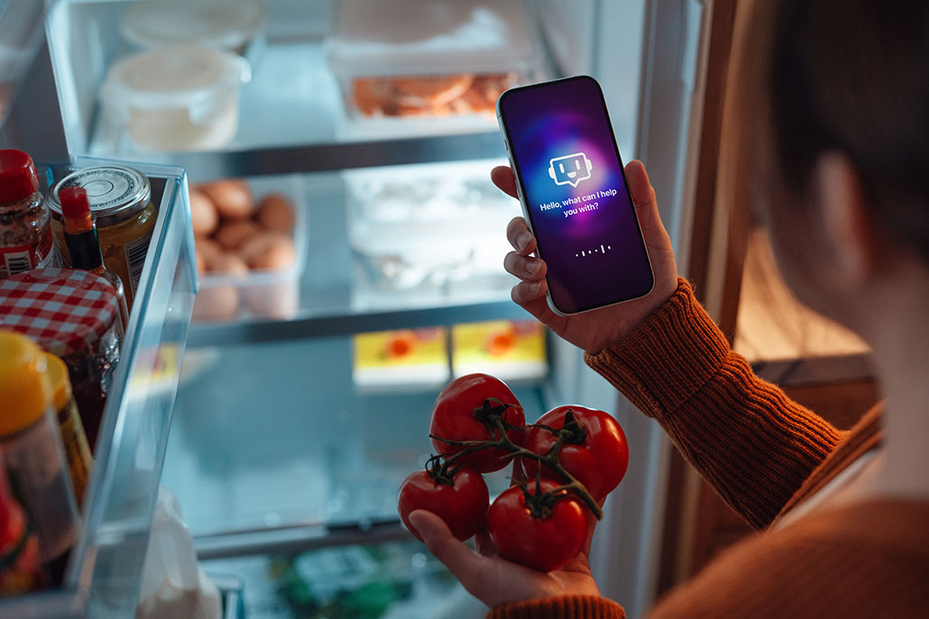 A person holds a smartphone displaying an AI chatbot interface while selecting tomatoes from an open refrigerator at night.
