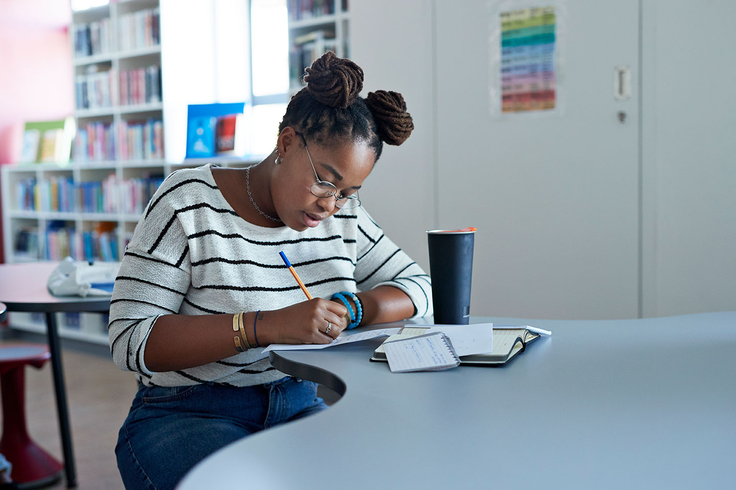 a young Black woman with space buns leans over a desk in a study center writing