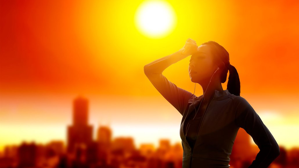 A heated woman wearing earbuds shields her eyes from an intense sun while standing outdoors silhouetted against a vivid orange sky, with a city skyline visible in the background.