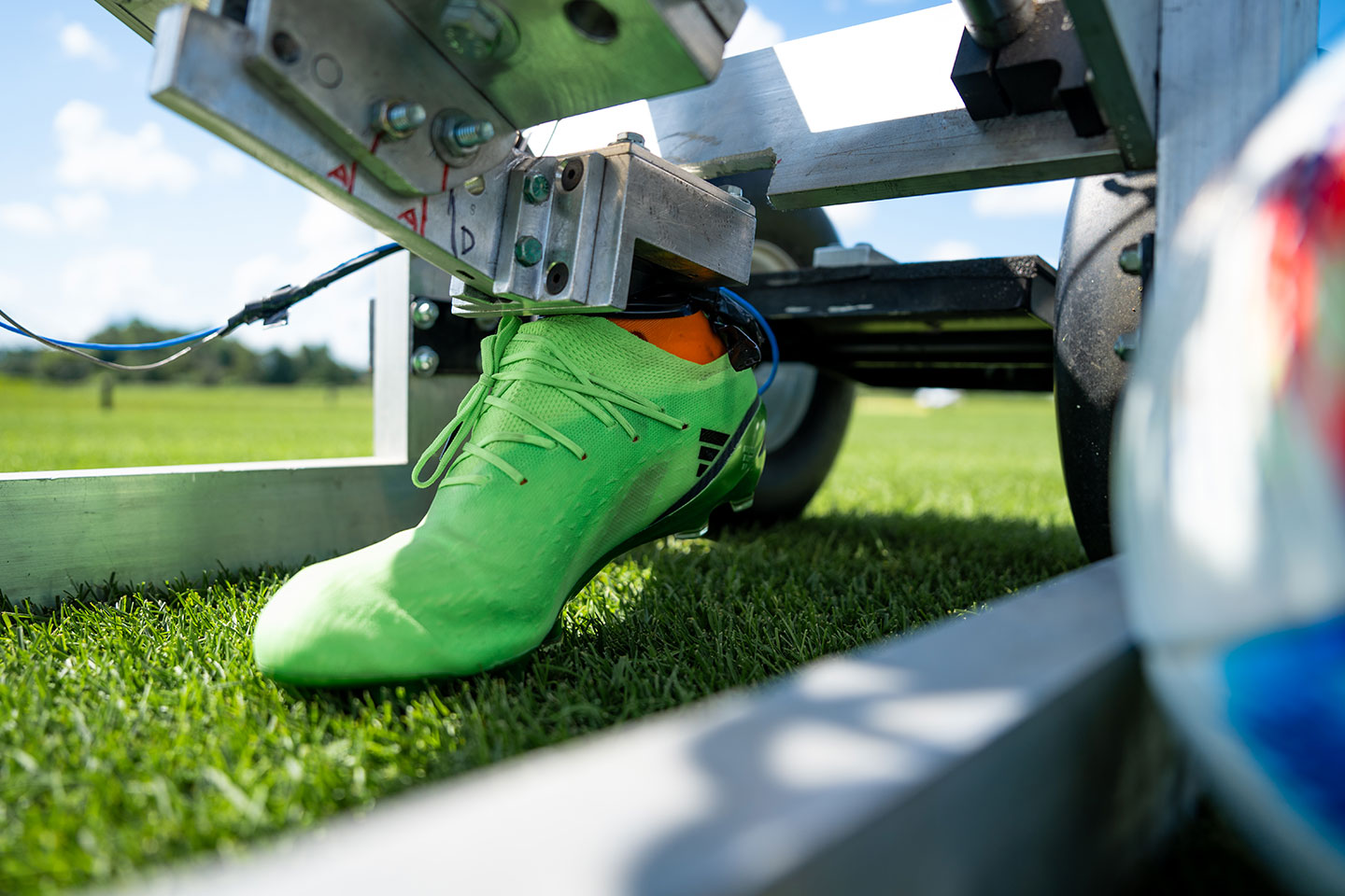 A bright green soccer cleat mounted to a mechanical testing device presses into a grass surface outdoors.