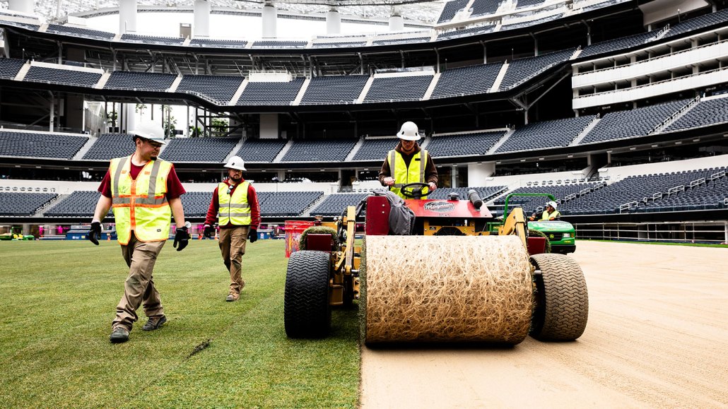 Three workers in hard hats and high-visibility vests walk alongside a machine rolling out sod inside an empty stadium.