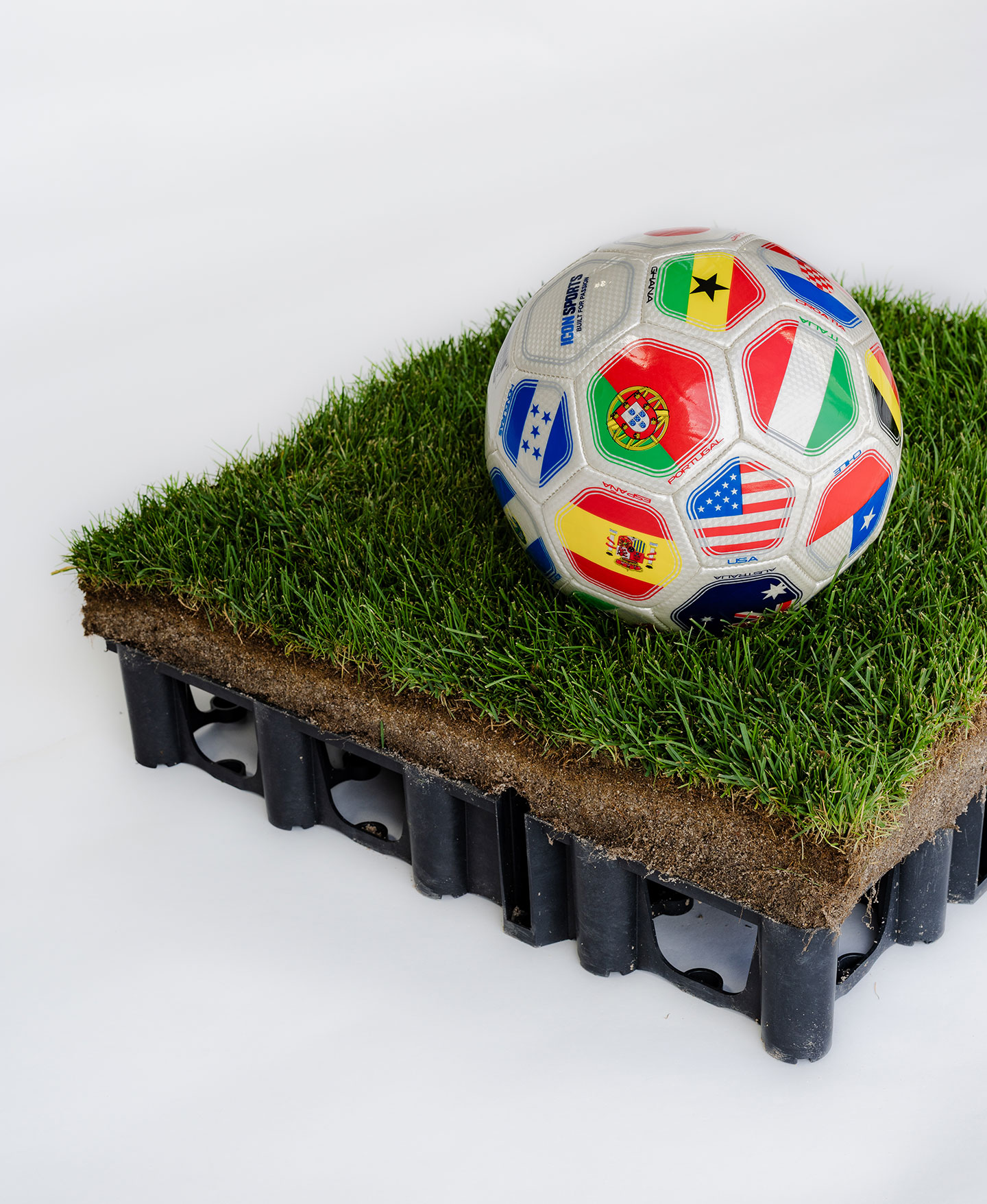 A soccer ball printed with international flags rests on a small square of sod sitting on a black plastic drainage tray, against a white background.