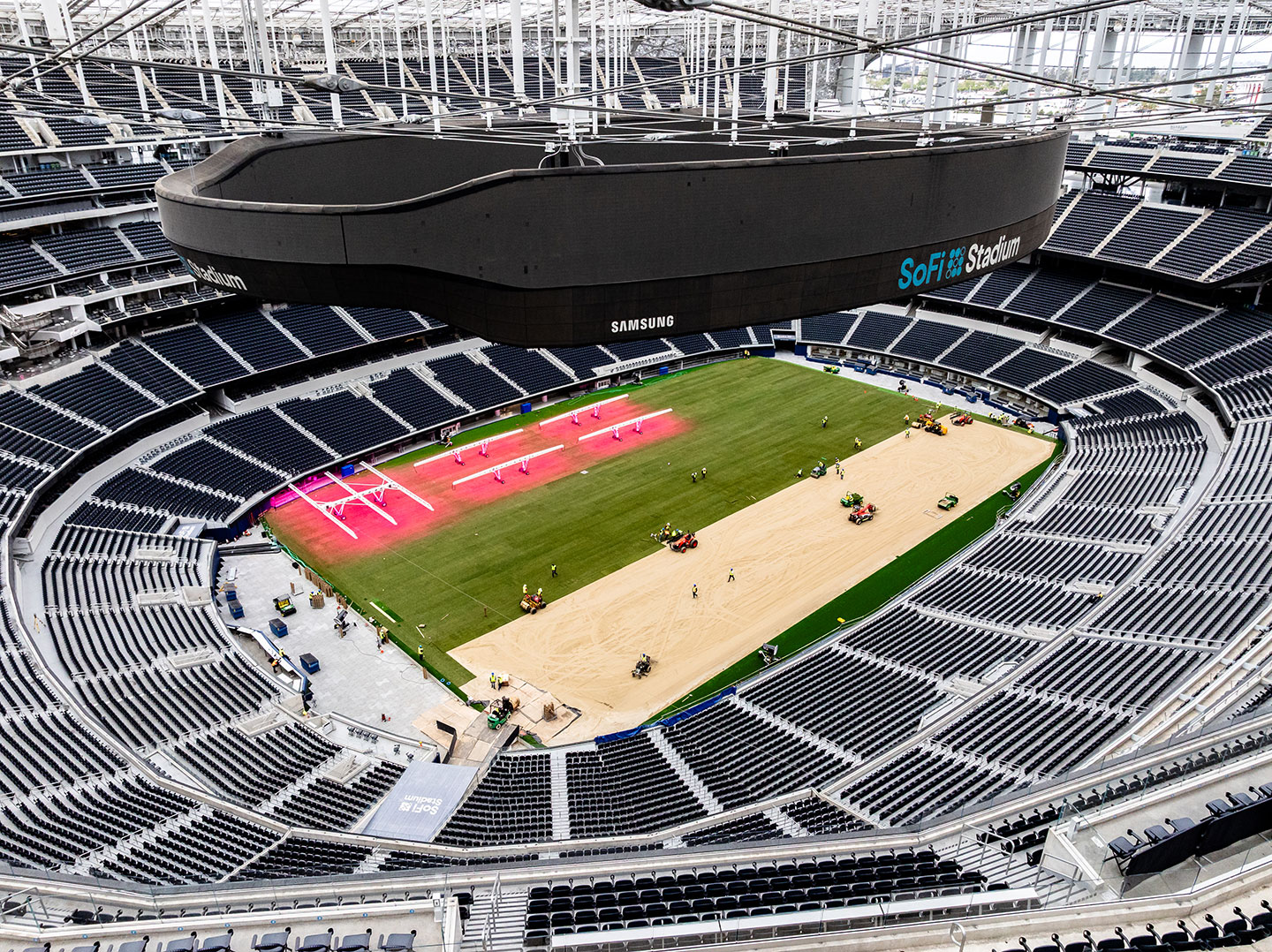 An aerial view of SoFi Stadium showing the field mid-installation, with sod laid on one half and a sandy base exposed on the other, surrounded by pink grow lights and workers on machinery.