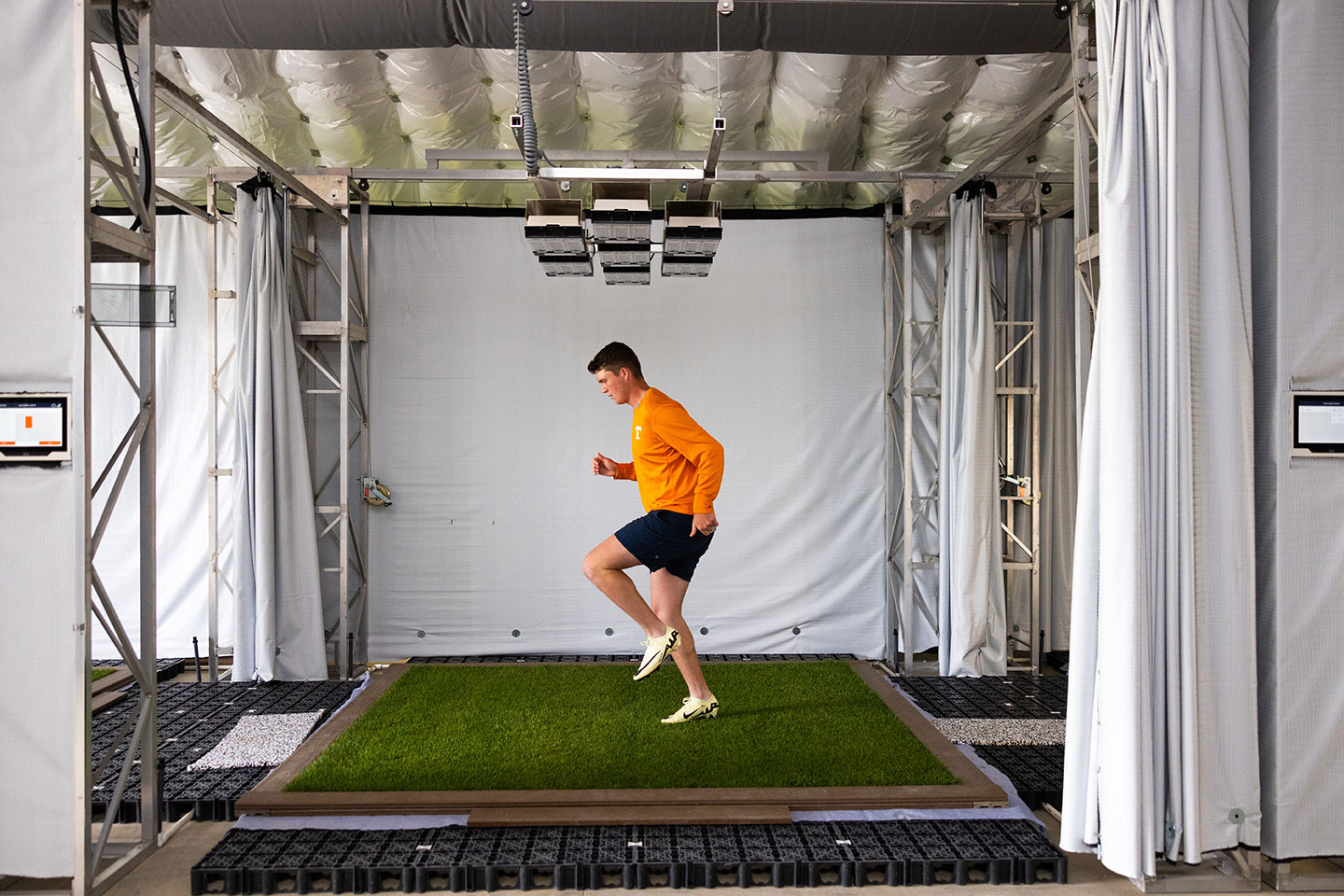 A person in an orange sweatshirt and cleats runs on a small patch of grass inside an enclosed testing facility with white curtains and overhead equipment.