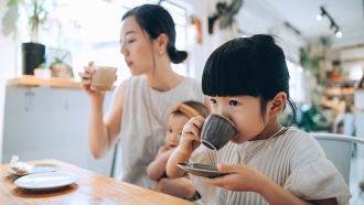 a little girl drinks a babycino at a cafe, she is sitting with her mom (also drinking coffee) who is holding a baby