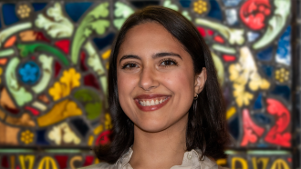 a photo of a smiling woman from the shoulders up. She has tan skin and brown hair and eyes standing in front of a colorful background