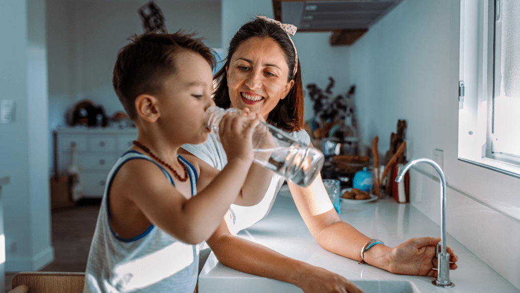 a young boy drinks from a glass of tap water, standing at the kitchen sink with his mom