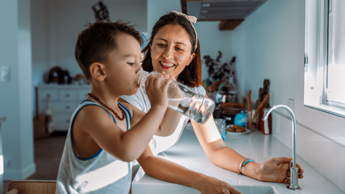 a young boy drinks from a glass of tap water, standing at the kitchen sink with his mom