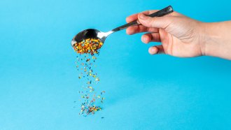 a hand holds a spoon pouring out a stream of colored microplastics on a blue background