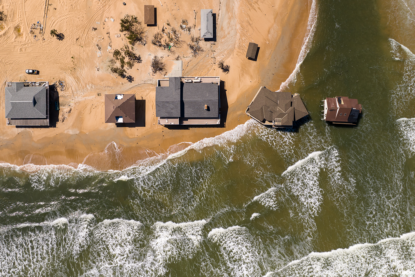 An aerial view of houses on a beach. Two of the house are partly or completely in the water.