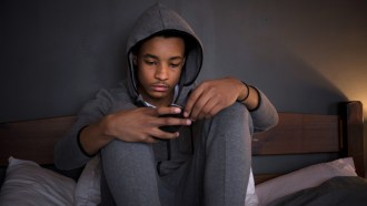 A teenager in a gray sweatshirt and sweatpants with a sad expression sits on a bed looking at a smartphone screen