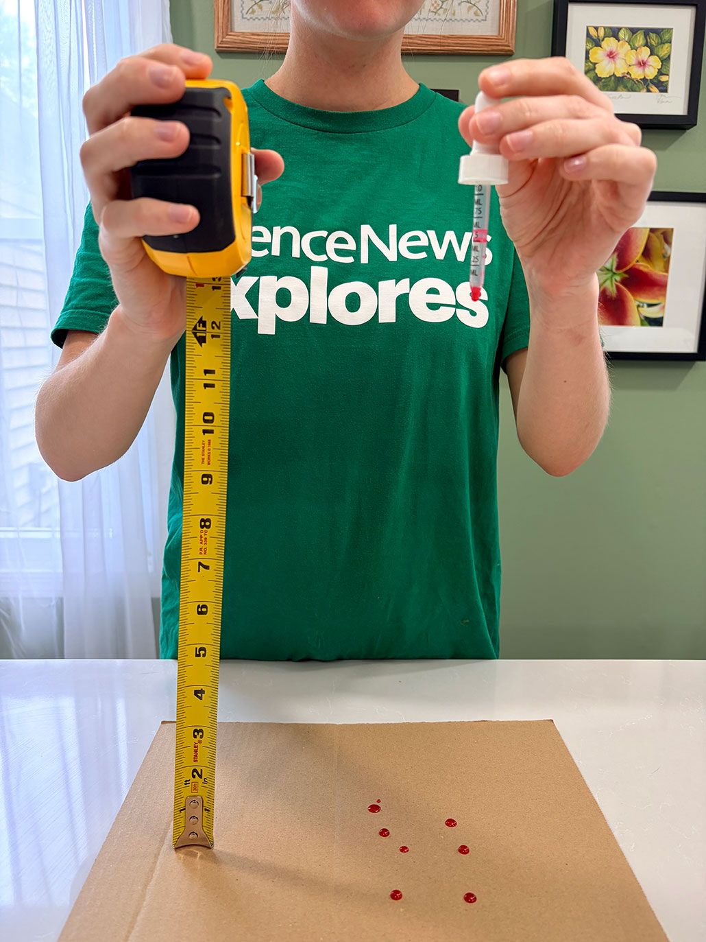 a person holds a tape measure vertically 12 inches high, and a dropper filled with red liquid on the same level, both over a piece of cardboard