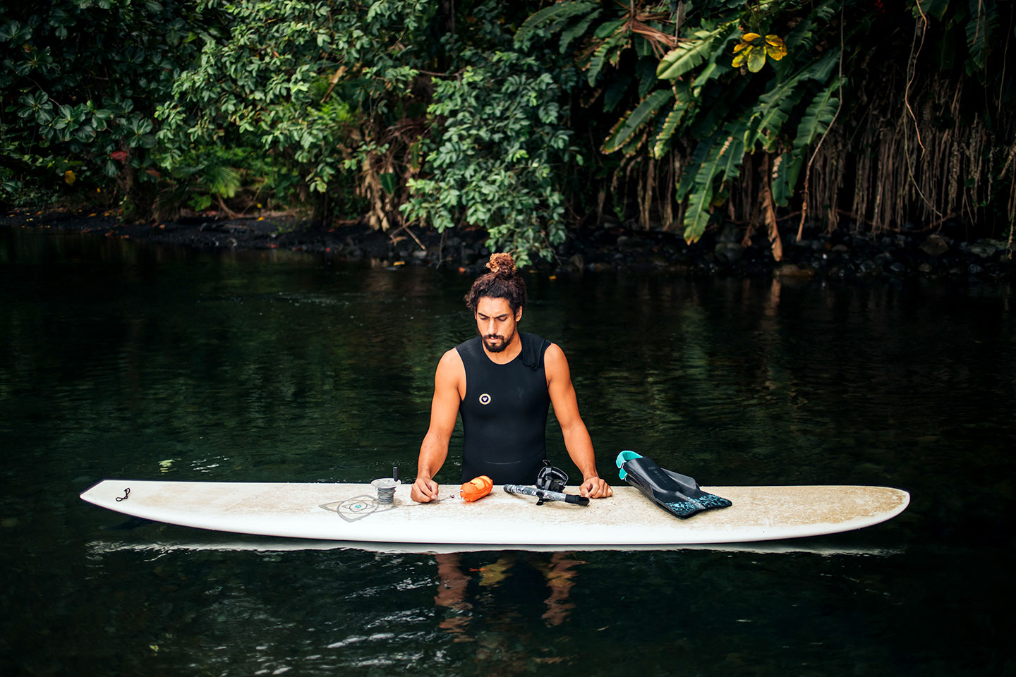 A person in a black sleeveless wetsuit standing in dark water, leaning over a surfboard. The board holds fins, a snorkel, and a small orange cylinder. Thick green plants fill the background.