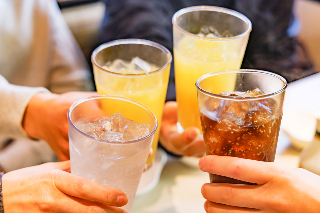 hands holding drinks on a table, juice, clear soda and a dark cola soda