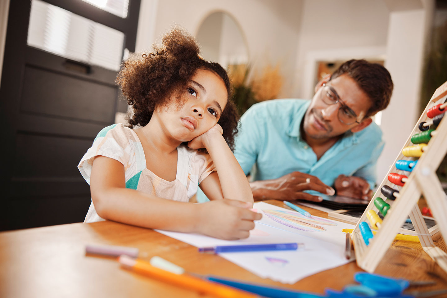 a young girl sits at a table doing homework but she is distracted and staring off into space