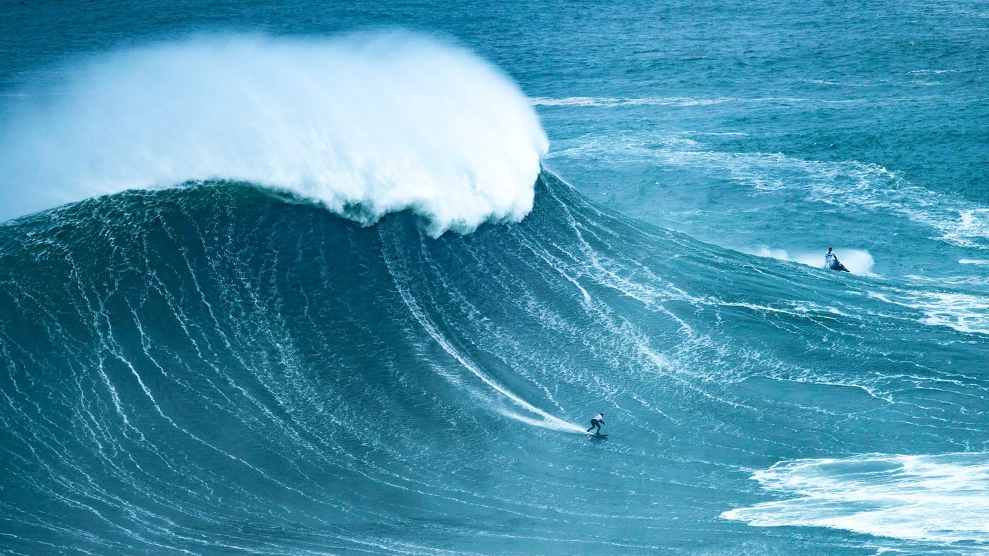 A giant wave crests as a very small person surfs across it